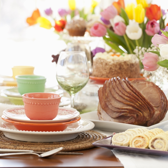 Table set for easter dinner with colorful plates, a spiral ham, carrot cake, pinwheel appetizers, and colorful tulips